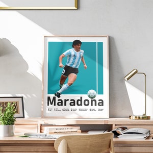 May include: A framed poster of Diego Maradona in his Argentina kit, set against a teal backdrop. The poster includes the name "Maradona" and a list of his achievements. The poster is displayed on a wooden desk, with a gold desk lamp.