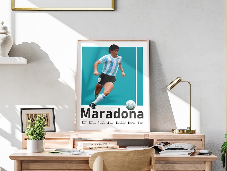 May include: A framed poster of Diego Maradona in his Argentina kit, set against a teal backdrop. The poster includes the name "Maradona" and a list of his achievements. The poster is displayed on a wooden desk, with a gold desk lamp.