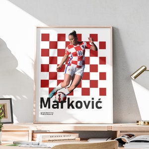 May include: A poster featuring a female soccer player in a red and white checkered uniform, kicking a soccer ball. The player's name, "Marković", is written in large, bold letters below the image. The text "1.  Senior Women's Cup 2021-22 Runner-Up" is written in smaller font below the name.