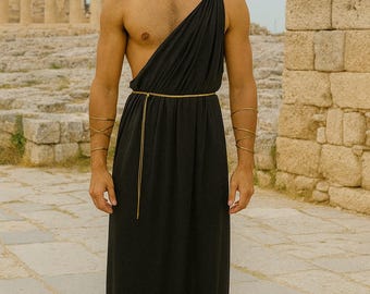 May include: A black toga-style dress with a gold waist belt and shoulder clasp. The model wears gold arm cuffs and a gold laurel wreath. Brown leather sandals complete the look. The background features ancient ruins.