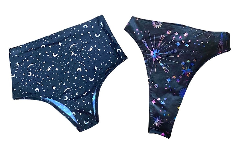 May include: Two pairs of underwear. One pair is navy blue with a white star and crescent moon pattern. The other pair is black with a colorful, iridescent starburst and star pattern.