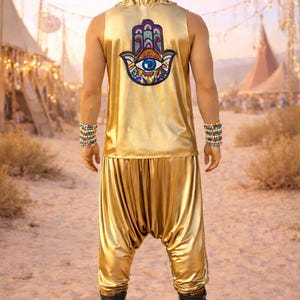 May include: Gold hooded sleeveless top and harem pants set. The top features a colorful Hamsa hand patch with an eye design. The outfit is paired with black boots and colorful beaded bracelets. The background shows a desert landscape.