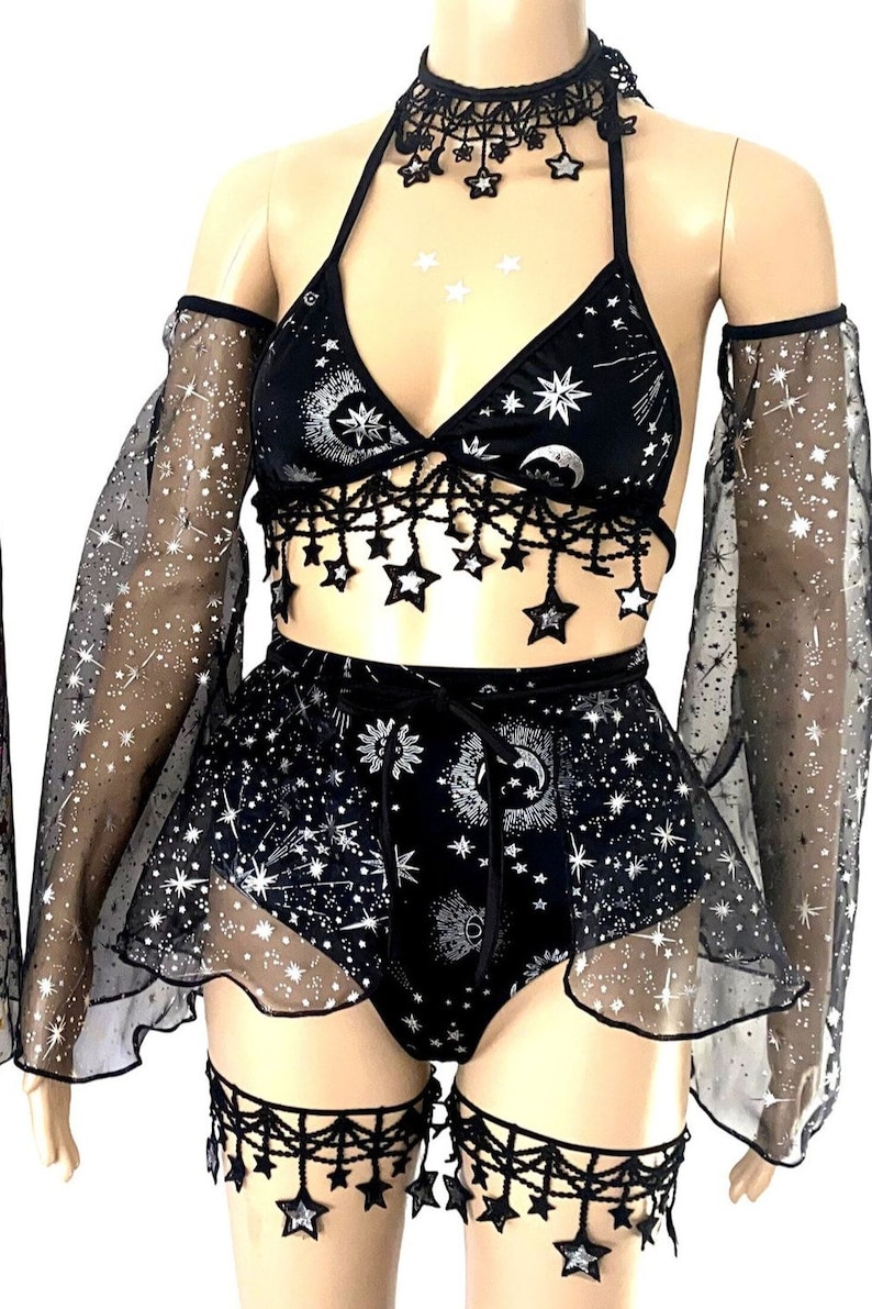 May include: Black celestial-themed lingerie set. The set includes a halter top, shorts, arm sleeves, a choker, and leg garters. The fabric is black with a silver celestial print of moons, stars, and suns. The edges are trimmed with star-shaped fringe.