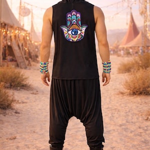 May include: Black hooded tank top and harem pants. The tank top features a colorful Hamsa hand design on the back. The person is wearing multiple beaded bracelets and black boots. The background shows a desert landscape with tents.