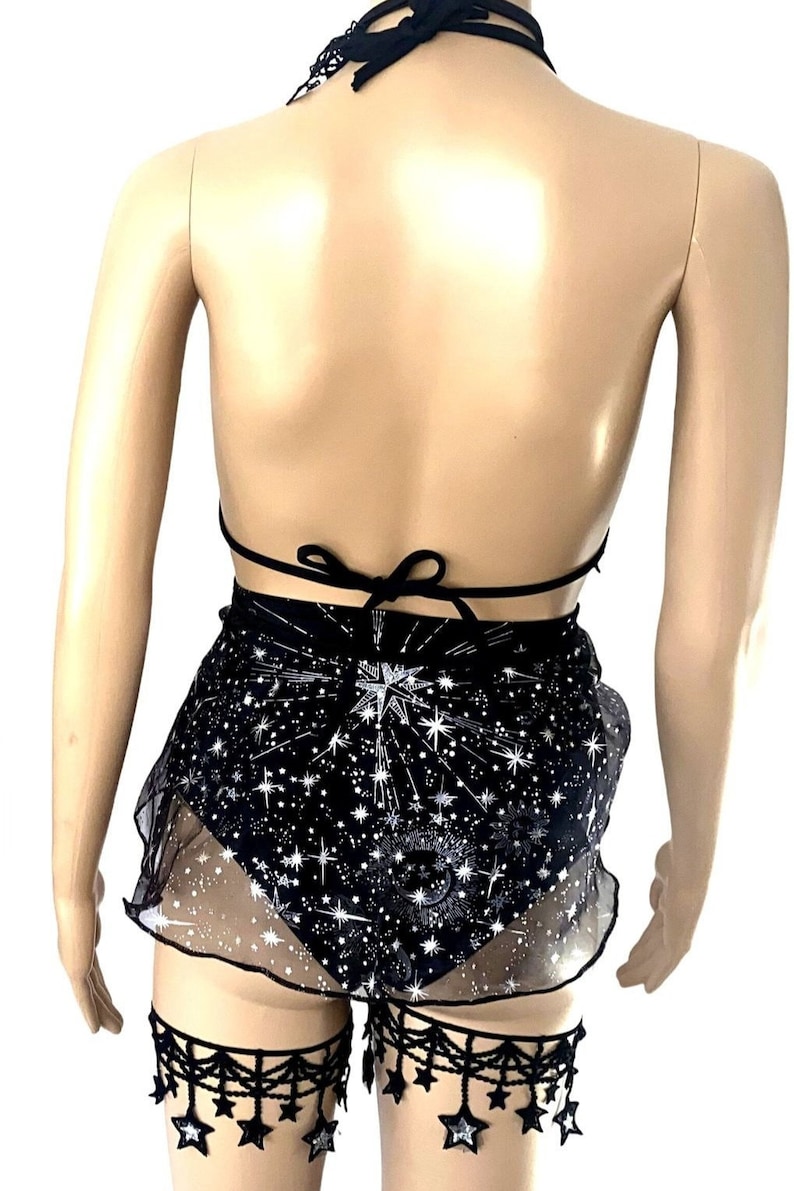 May include: Black halter top, shorts, and garter set. The top ties at the neck and back. The shorts are black with a white star pattern and a sheer overlay. Black star-shaped garter accents complete the set. A celestial-themed outfit.