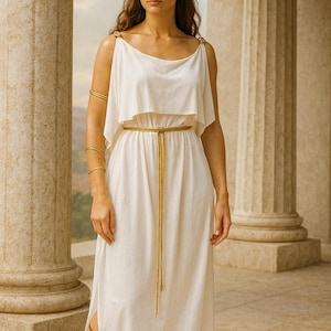 May include: A white Grecian-style dress with a gold belt and shoulder accents. The dress has a flowing design and is paired with gold arm cuffs and brown sandals. A floral crown adorns the head. The setting is an outdoor location with columns.