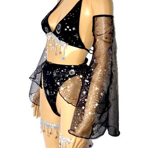 May include: Black lingerie set with a celestial theme. The set includes a halter top, high-waisted bottoms, arm sleeves, a choker, and leg garters. The fabric is black with a starry pattern and silver accents. The set is embellished with dangling star charms.