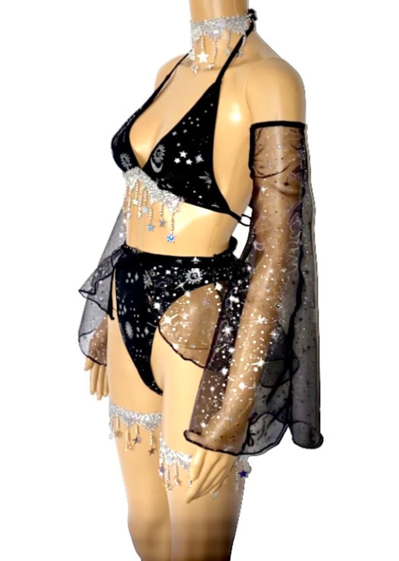 May include: Black lingerie set with a celestial theme. The set includes a halter top, high-waisted bottoms, arm sleeves, a choker, and leg garters. The fabric is black with a starry pattern and silver accents. The set is embellished with dangling star charms.