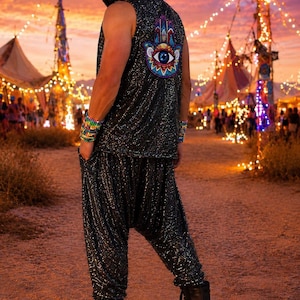 Puede incluir: Una camiseta sin mangas con capucha y pantalones holgados a juego, ambos de color negro con un acabado brillante y reluciente. La parte posterior de la camiseta presenta un gran parche bordado y colorido de una mano de Hamsa con un ojo azul.