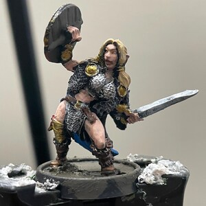 May include: A hand-painted miniature figurine of a warrior with blonde hair, wearing silver chainmail armor, gold accents, and brown boots. The figure holds a round shield raised in the right hand and a silver sword extended in the left hand.
