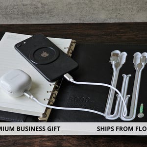 May include: A black smartphone with a MagSafe charger is connected to a white charging cable, resting on a notebook. A white earbud case is also connected to a charging cable. Additional white charging cables are visible. The image is labeled "PREMIUM BUSINESS GIFT".