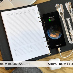 May include: A black folio organizer featuring a ring-bound planner with lined pages, a smartphone displaying a 22% charge, and integrated charging cables. The organizer includes a dedicated slot for a white wireless earbud charging case.
