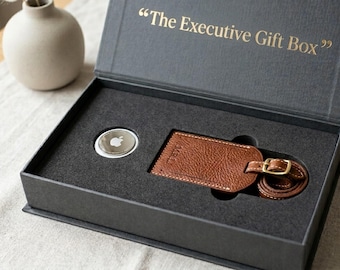 May include: A black rectangular gift box with the text The Executive Gift Box printed in gold on the inside lid. The box contains a silver circular tracking device with an Apple logo and a brown textured leather luggage tag with a gold buckle.