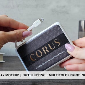 May include: A white and black power bank with the word "CORUS" in gold lettering. The power bank is connected to a white charging cable. The text "Creating Harmony between Faith & Finance" is also visible.