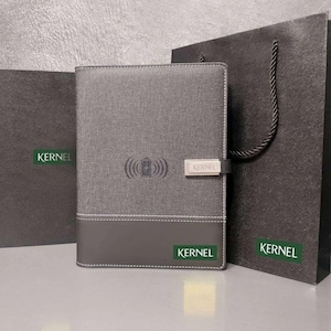 May include: A gray fabric-covered notebook with a dark gray bottom panel and white stitching. The cover features a wireless charging icon and a silver metal clasp labeled KERNEL. It is displayed alongside a black gift bag with the KERNEL logo.