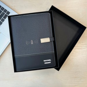 May include: A dark gray and black notebook with a wireless charging icon and a gold-toned metal clasp. The cover features the name RACHEL GRANT in white text. The notebook is presented inside a black rectangular gift box with a matching lid.
