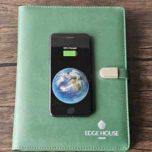 May include: A sage green leather-like notebook with a silver metal clasp closure and the white printed text "EDGE HOUSE MIAMI" sits beneath a black smartphone displaying an 85% battery charge icon and a circular image of the Earth on its screen.