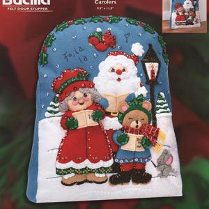 May include: A blue felt door stopper kit titled Carolers by Bucilla, measuring 9.5 inches by 11.5 inches. The design features Santa Claus, a woman, and a bear singing carols with a street lamp, a mouse, and the text Fa, la, la.