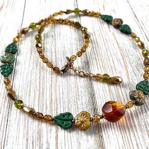 May include: A beaded necklace featuring a mix of amber-colored faceted beads, small green glass beads, and dark green leaf-shaped and flower-shaped beads. The necklace includes a metallic clasp and an adjustable chain extension.