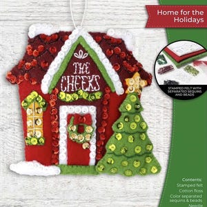 May include: Felt ornament kit featuring a red house with white trim, a green roof, and a Christmas tree. The house has the text "THE CHEEKS" on the front. Includes stamped felt, sequins, beads, cotton floss, and a needle. Measures 4.5 inches by 4.5 inches.