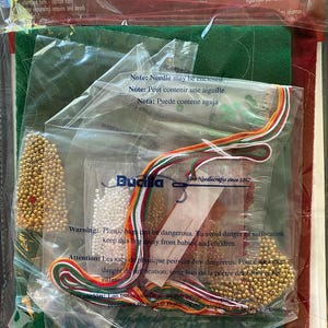 May include: A Bucilla craft kit in a clear plastic bag. The kit includes felt, floss, beads, and sequins in various colors, including green, red, and gold. The packaging has text in English, French, and Spanish, with a warning about plastic bags.