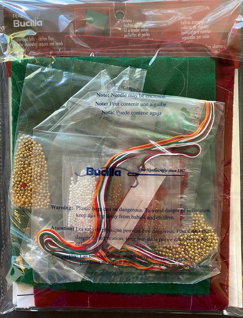 May include: A Bucilla craft kit in a clear plastic bag. The kit includes felt, floss, beads, and sequins in various colors, including green, red, and gold. The packaging has text in English, French, and Spanish, with a warning about plastic bags.