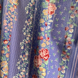 May include: Close-up of a floral fabric with a blue background. The fabric features vertical stripes and floral patterns with roses, daisies, and other flowers in shades of pink, yellow, and white. The fabric has a vintage aesthetic.