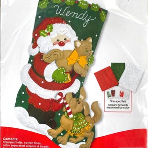 May include: A Bucilla Santa's Best Friends felt stocking kit measuring 18 inches. The green stocking features a Santa Claus holding a tan cat, a dog with a candy cane, and the name Wendy embroidered in white. Includes felt, floss, and beads.