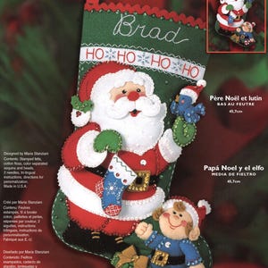 May include: A felt stocking kit featuring Santa Claus and an elf. The stocking is green with a red top and a design of Santa holding a blue bird and an elf. The text "Brad" and "HO HO HO" are embroidered on the stocking. The stocking is 18 inches.