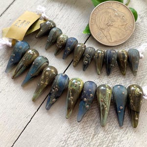 May include: Rows of teardrop-shaped beads in shades of blue, green, and brown, with a speckled design. A U.S. quarter coin is next to the beads for size comparison. A tag with the text "2014 Spike 1000" is attached to the beads.