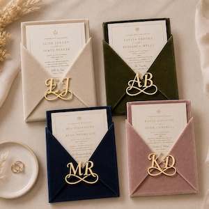 May include: Four velvet wedding invitation envelopes in beige, dark green, navy blue, and dusty rose. Each envelope features a gold-toned metal monogram clasp with initials EJ, AB, MR, and KD, holding a cream-colored card with printed black text.