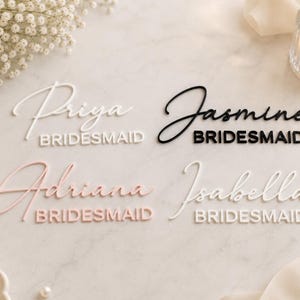 May include: Four custom name signs featuring the names Priya, Jasmine, Adriana, and Isabella in a script font, each paired with the word BRIDESMAID in a block font. The signs are rendered in white, black, and soft pink acrylic materials.