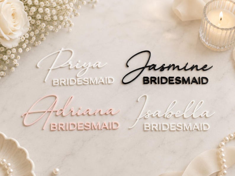 May include: Four custom name signs featuring the names Priya, Jasmine, Adriana, and Isabella in a script font, each paired with the word BRIDESMAID in a block font. The signs are rendered in white, black, and soft pink acrylic materials.