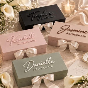 May include: Four rectangular gift boxes in black, light pink, dusty rose, and sage green, each featuring a satin ribbon bow and raised script lettering reading Taylor, Kendall, Jasmine, and Danielle, with the word BRIDESMAID printed below.
