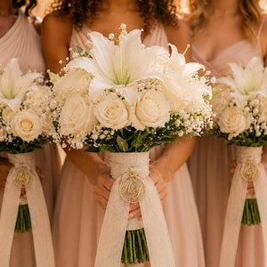 May include: Three floral bouquets featuring white lilies, cream roses, and baby's breath. Each bouquet is wrapped in a wide, textured beige ribbon with a circular gold charm engraved with the letters A&Y attached to the stems.