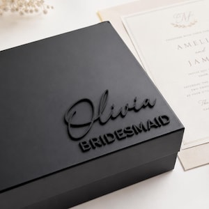 May include: A matte black rectangular gift box featuring the name Olivia in a raised script font and the word BRIDESMAID in a raised sans-serif font on the lid. The box is positioned next to a cream-colored wedding invitation card.