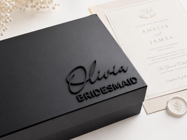 May include: A matte black rectangular gift box featuring the name Olivia in a raised script font and the word BRIDESMAID in a raised sans-serif font on the lid. The box is positioned next to a cream-colored wedding invitation card.