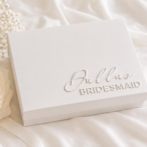 Puede incluir: Una caja de regalo rectangular blanca con el nombre Dallas en una fuente cursiva en relieve sobre la palabra BRIDESMAID en una fuente sans-serif en relieve. La caja está junto a una rosa blanca y flores pequeñas sobre tela blanca.