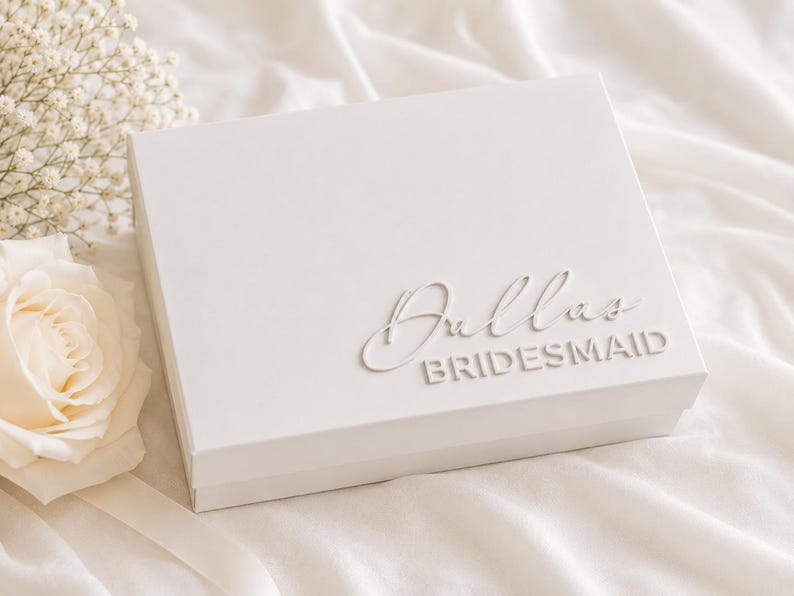 May include: A rectangular white gift box featuring the name Dallas in a raised script font above the word BRIDESMAID in a raised sans-serif font. The box is positioned next to a cream-colored rose and a sprig of white baby's breath flowers.