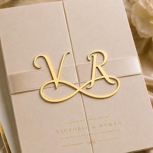 May include: A beige gatefold wedding invitation card featuring a gold infinity-shaped monogram with the letters V and R. The card includes the printed text Victoria & Roman, invite you to celebrate their marriage, and a horizontal satin ribbon.