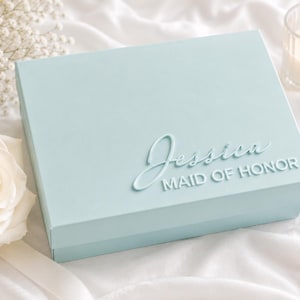 May include: A light blue rectangular gift box featuring the name Jessica in a raised script font and the words MAID OF HONOR in a raised sans-serif font on the lid. The box has a smooth matte finish and a clean, structured design.