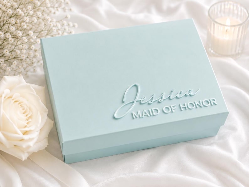 May include: A light blue rectangular gift box featuring the name Jessica in a raised script font and the words MAID OF HONOR in a raised sans-serif font on the lid. The box has a smooth matte finish and a clean, structured design.