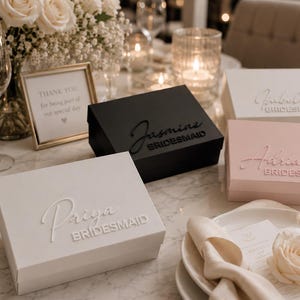 May include: Four rectangular gift boxes in white, black, and light pink, each featuring a raised script name and the word BRIDESMAID printed below. The boxes are arranged on a table with place settings, a floral arrangement, and a sign.