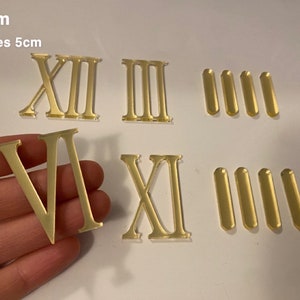 以下が含まれることがあります： 金色ののアクリル製ローマ数字と長方形のダッシュのセット。数字にはXII、III、VI、XIが含まれています。6cmおよびdashes 5cmというテキストが表示されています。数字の高さは6cm、ダッシュの長さは5cmです。