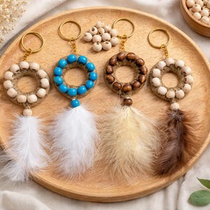 May include: Four handmade keychains with circular beaded designs in beige, blue, and brown, each adorned with a feather. The keychains are arranged on a wooden tray, alongside small bowls of beads and dried flowers. A natural, bohemian aesthetic.