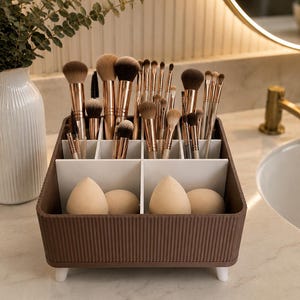 May include: A brown, rectangular makeup organizer with a ribbed exterior and four small white feet. The interior features white dividers creating multiple compartments holding various makeup brushes with rose gold ferrules and four beige makeup sponges.