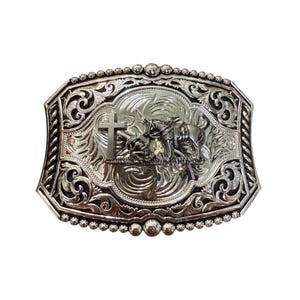 May include: Silver-toned rectangular belt buckle with an ornate design. The buckle features a central scene with a cross, a horse, and a rider, surrounded by detailed scrollwork and a beaded border. A classic Western accessory.