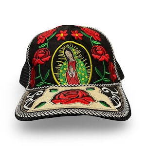 May include: A black trucker hat featuring an embroidered image of the Virgin of Guadalupe surrounded by red roses on the front panel. The brim displays a beige rectangular patch with a large red rose and decorative white stitching along the edges.