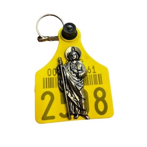 May include: Yellow plastic tag with a metal charm of a saint holding a staff. The tag has a barcode and the number "00 298 61" printed on it.