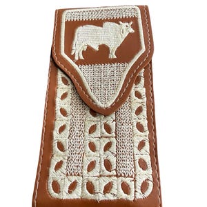 May include: A brown leather wallet with a white embroidered bull design on the flap. The wallet features intricate white stitching and a decorative pattern. The wallet is closed with a flap and is designed to hold cards and cash.